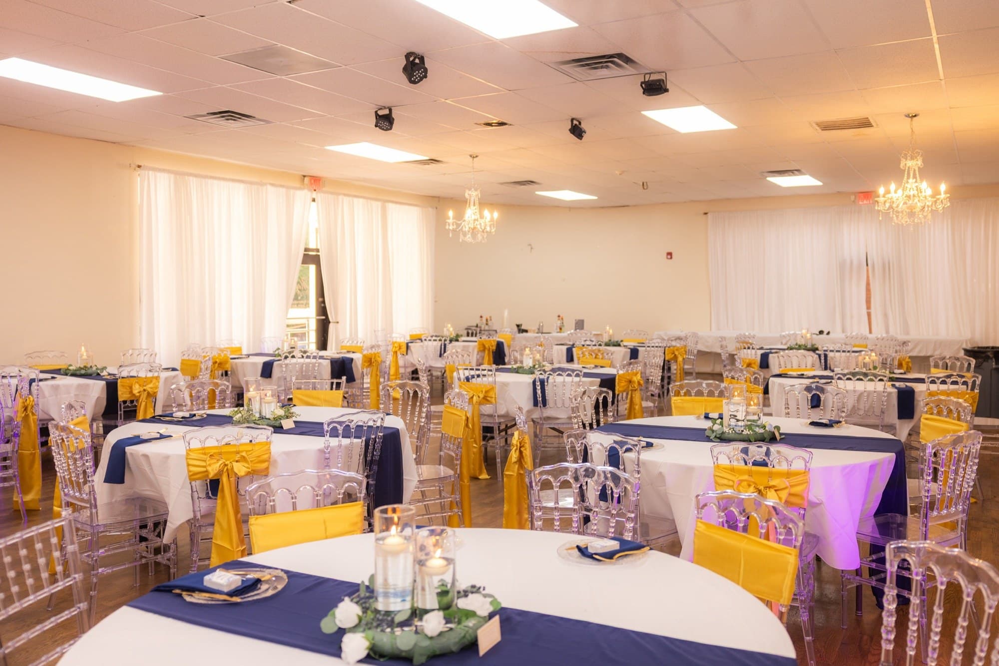 Salón de recepción con manteles azules, moños dorados y sillas de cristal