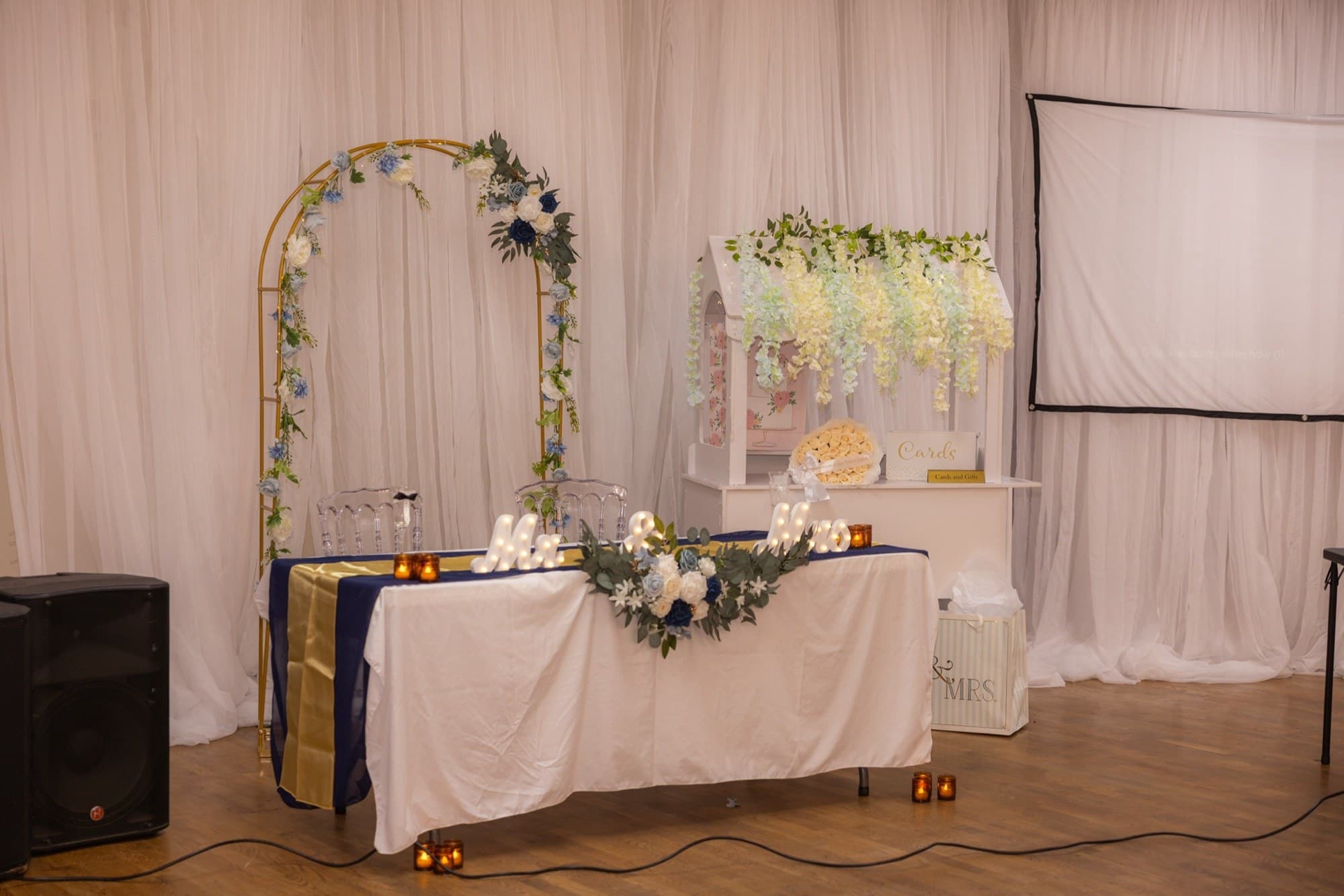 Mesa de novios bajo un arco floral dorado con casita para tarjetas