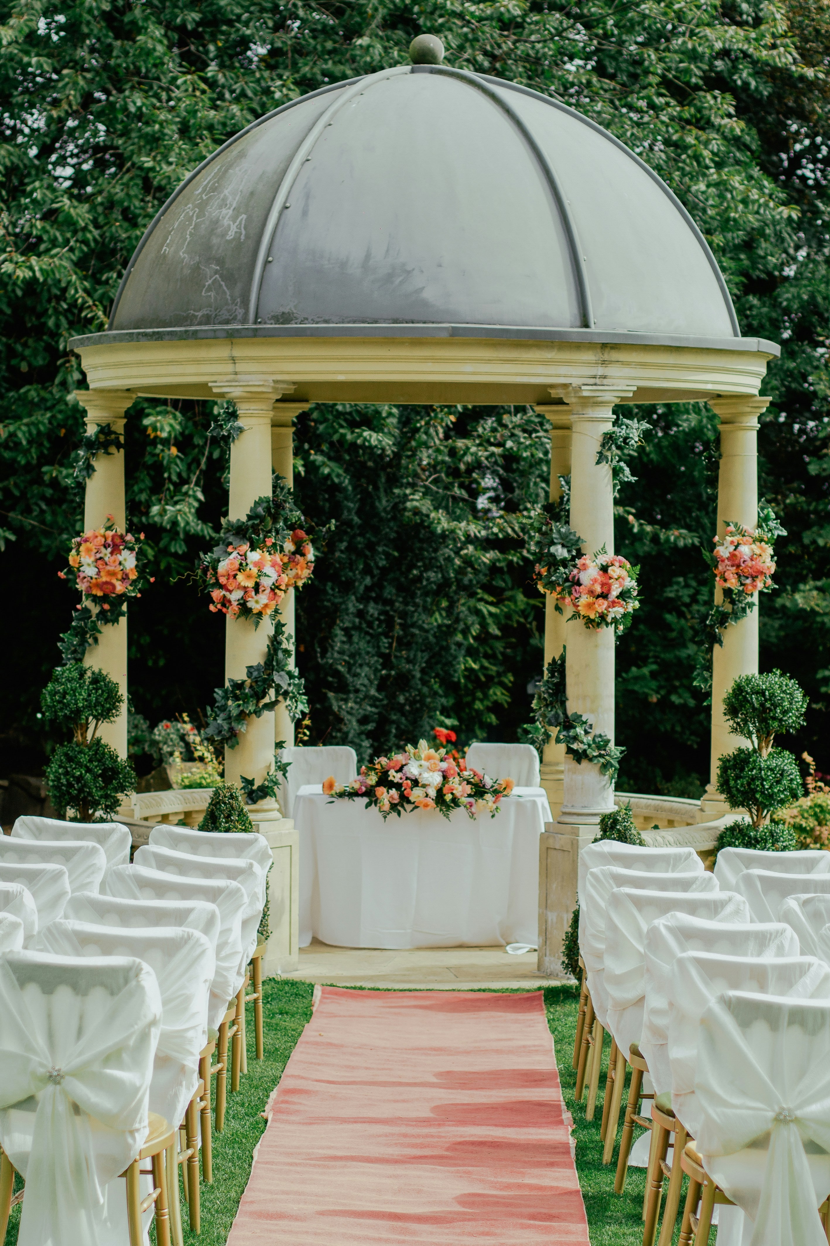 An elegant outdoor wedding ceremony with a floral-draped rotunda and a blush aisle runner