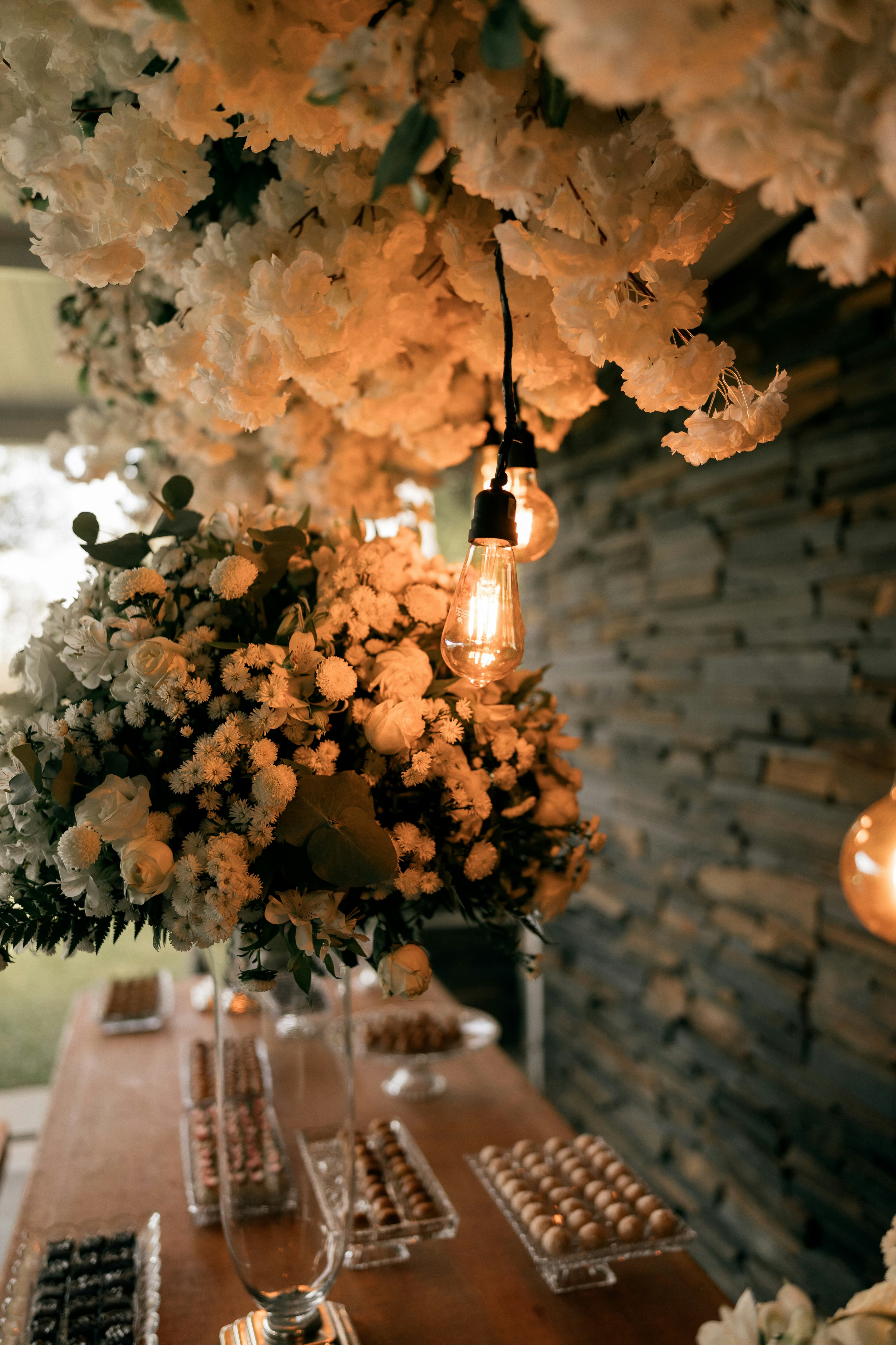 Lush white florals and warm Edison-bulb lighting above a styled dessert table