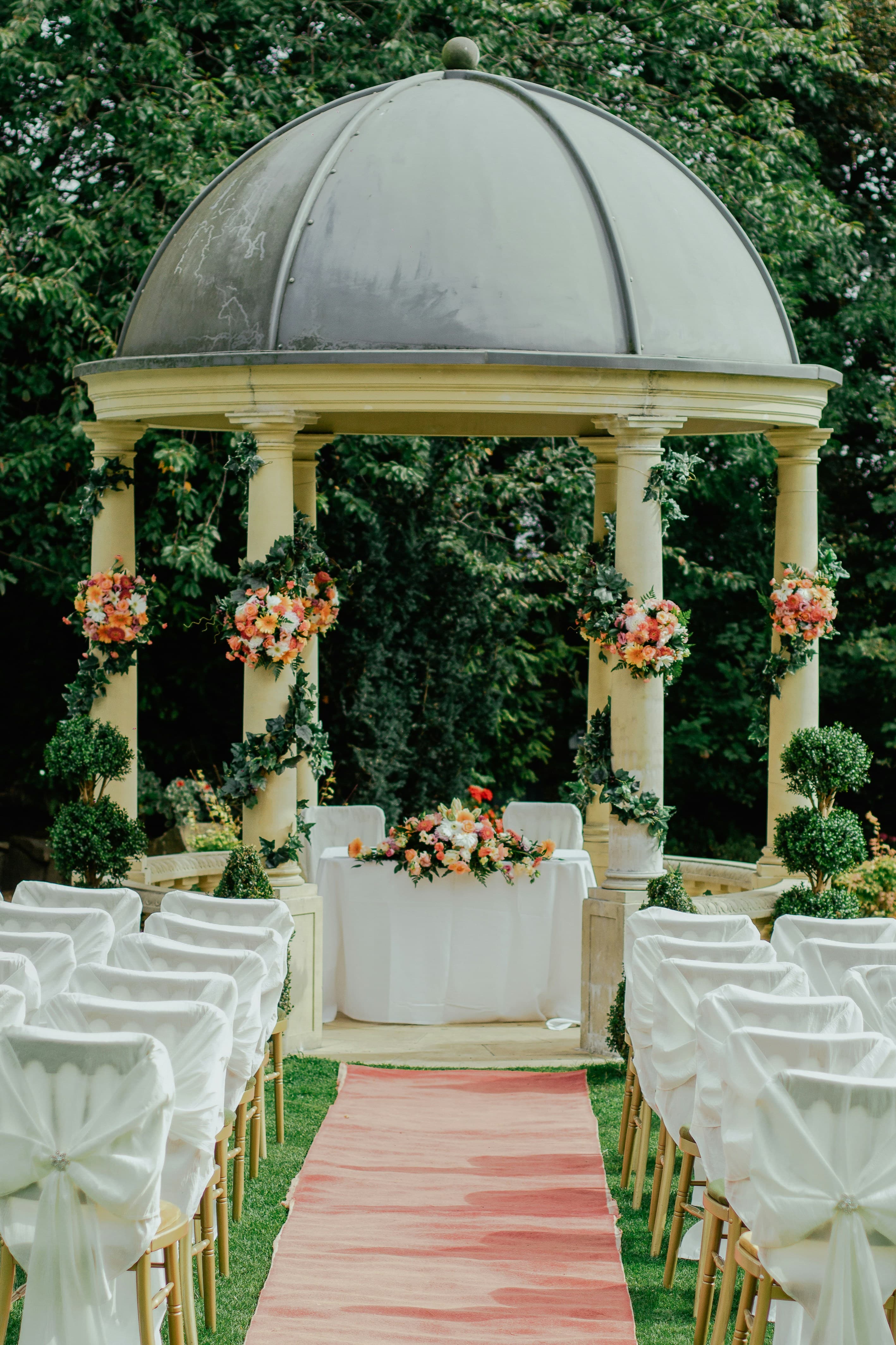 An elegant outdoor wedding ceremony with a floral-draped rotunda and a blush aisle runner